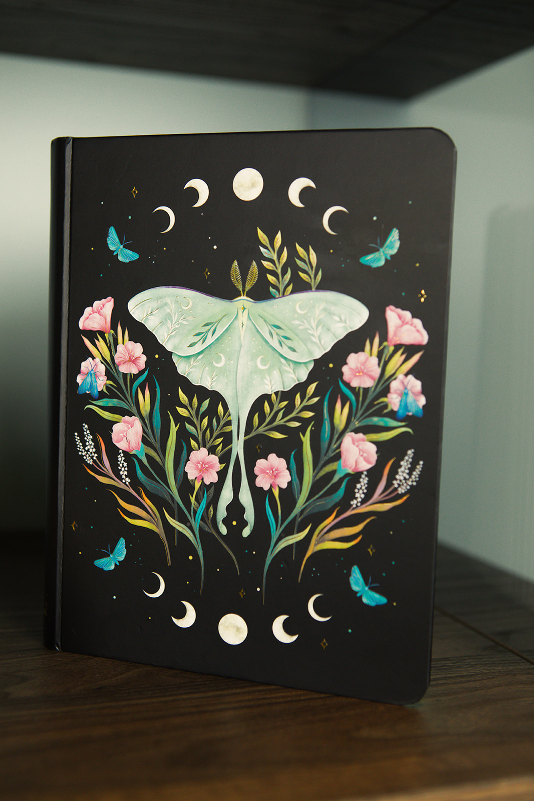 A journal with a luna moth and the phases of the moon painted on the cover, standing on a wooden shelf