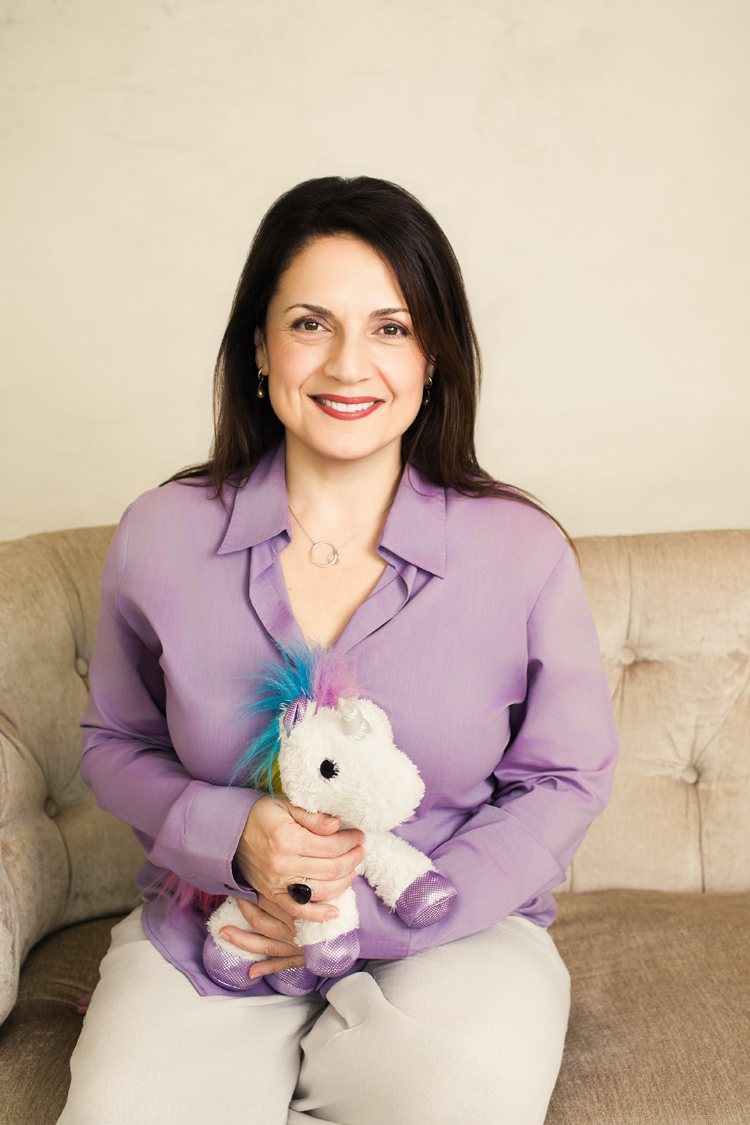 Nina seated on a sofa, smiling, holding a small stuffed unicorn
