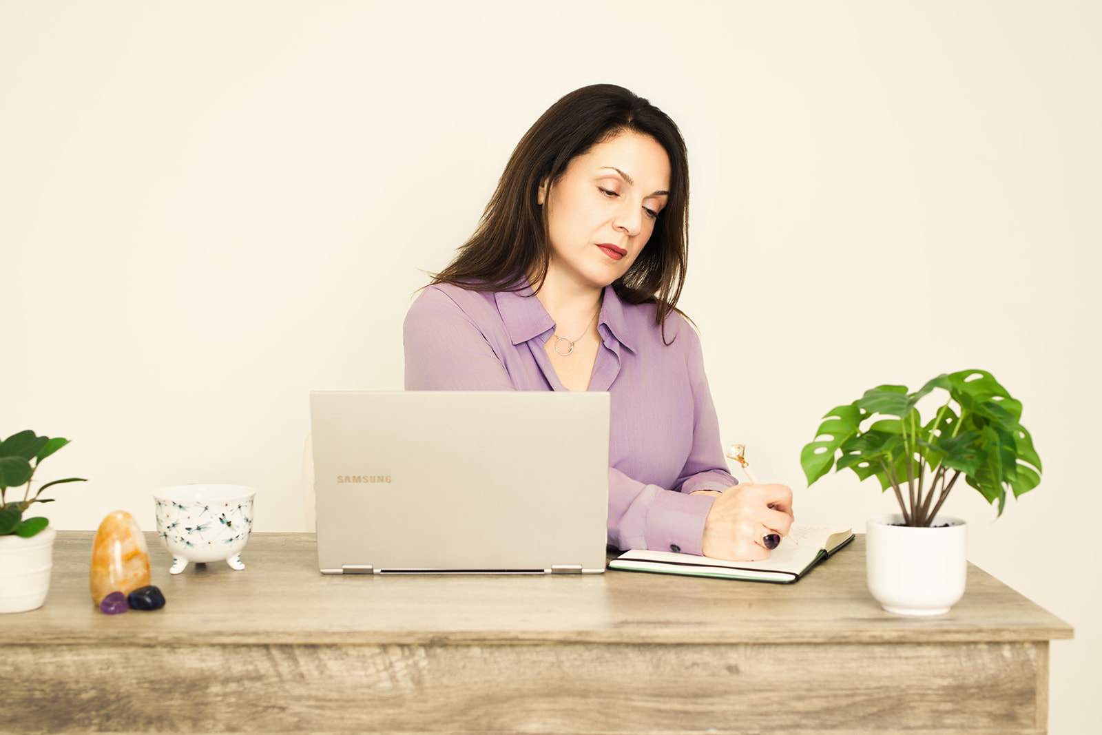 Nina writing in a notebook at her desk beside her laptop and a small plant